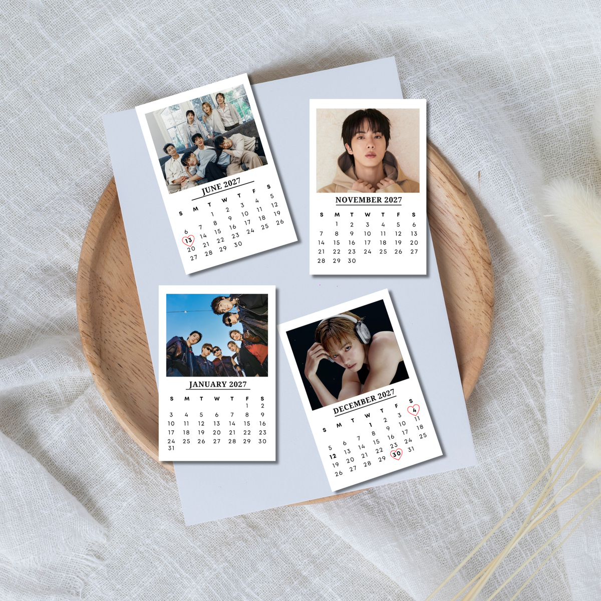 Four custom photo calendar cards for a year, displayed on a light blue sheet of paper over a round wooden tray. The cards feature placeholder spots for group and individual member photos, with the months June, November, January, and December.